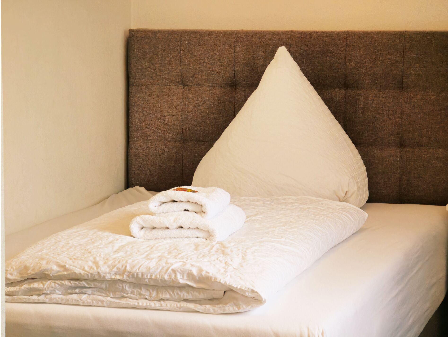 A neatly made bed with a brown headboard, white pillows, and a folded white towel.