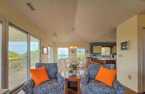 A spacious dining area with a large table, two patterned sofas with orange cushions, and a view of the outdoors. - Double Vision – Chill with Sound and Ocean Views! (Surf City)