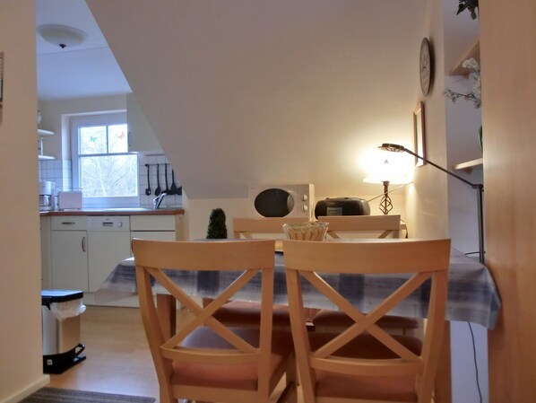 A dining area with a table and chairs, a lamp, a clock, and a kitchen in the background.