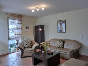 A living room with a sofa, armchair, coffee table, and a radiator. There is a window with curtains, a framed picture on the wall, and a lamp. - 2-zimmer-fewo,im 2.og für 2 Pers (Graal-Müritz)