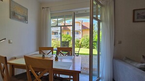A dining area with a table and chairs, a window with curtains, and a view of a building and greenery outside. - 2-zi-ferwo für 3 Pers im EG. Wlan (Graal-Müritz)