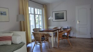 Une pièce avec un canapé blanc, des chaises en osier, une table en bois, une fenêtre avec des rideaux, un tableau encadré au mur et une porte.