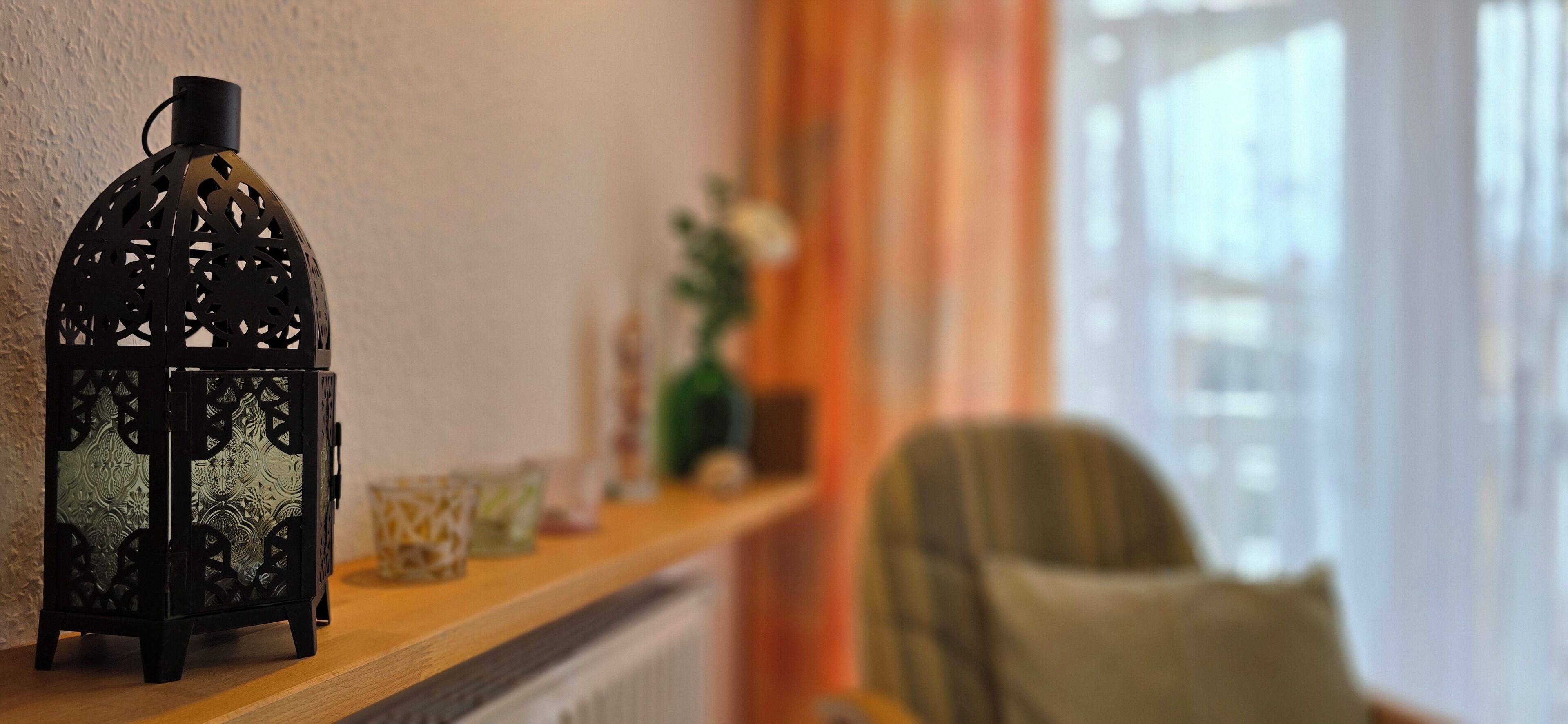 A room with a decorative black lantern on a wooden shelf, a chair, and a window with curtains.