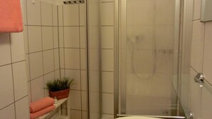 A bathroom with white tiles, a glass shower enclosure, a white bathtub, a pink towel, and a small potted plant on a shelf.