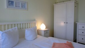 Una cama bien hecha con ropa de cama y almohadas blancas, una lámpara de noche, un armario y una mesita de noche pequeña con cajones.