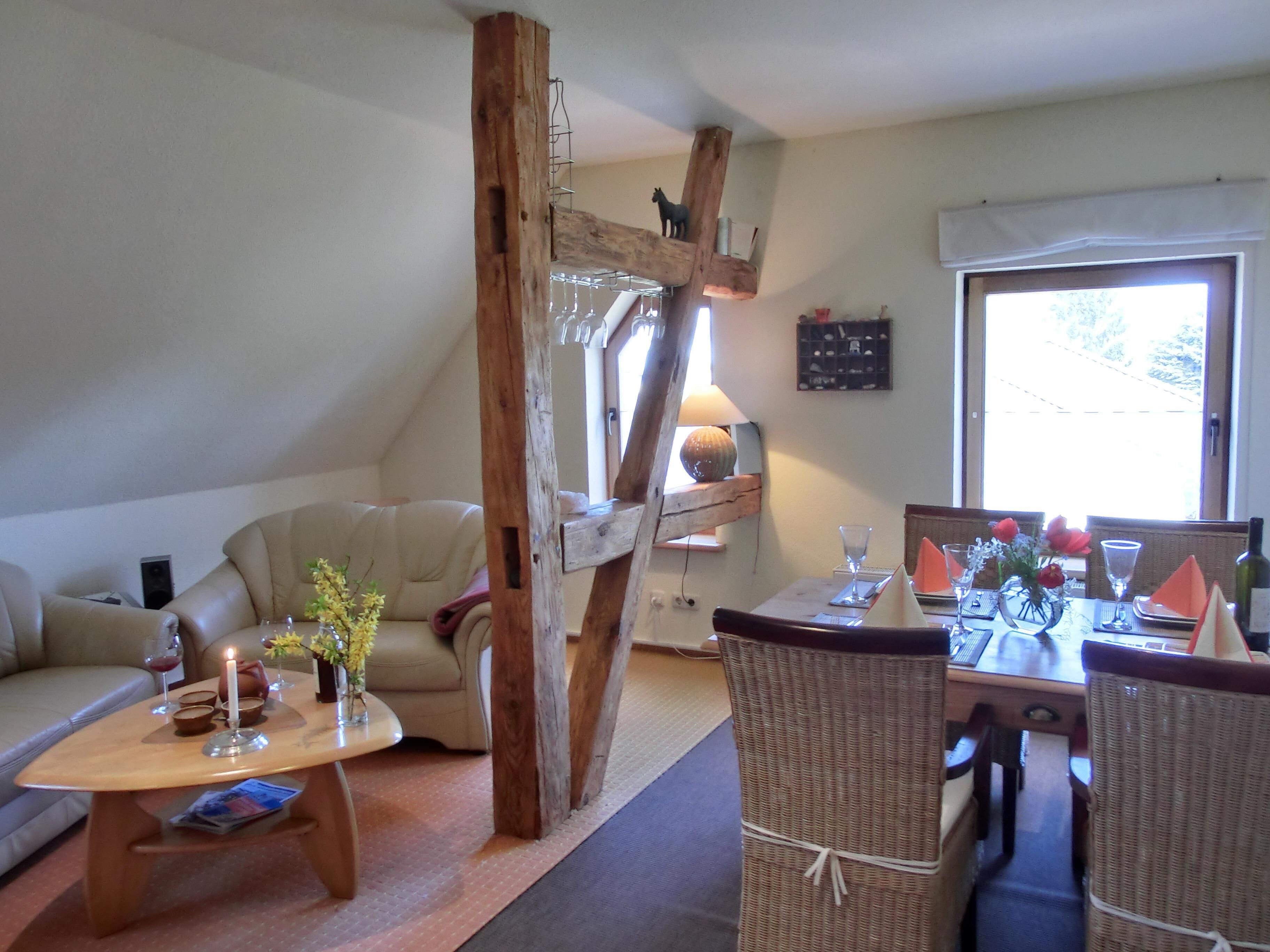 A cozy living room with a wooden beam structure, a sofa, a dining table set with orange napkins, and a wicker chair.