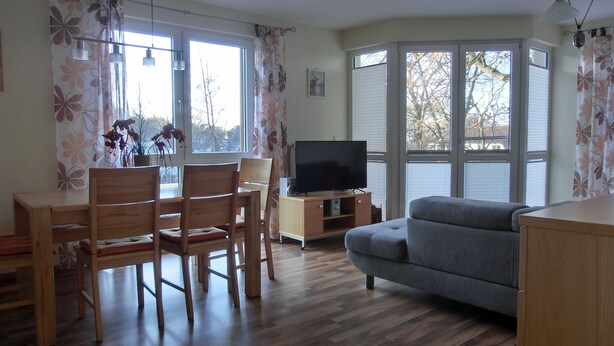 A living room with a wooden dining table, two chairs, a grey sofa, a TV on a wooden stand, and large windows with curtains. - 3-room apartment close to the beach for up to 6 people with elevator (Graal-Müritz)