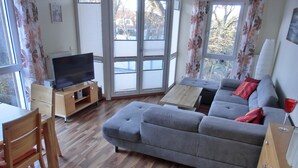 A living room with a grey sofa, wooden flooring, a TV on a wooden stand, and a window with curtains. - 3-room apartment close to the beach for up to 6 people with elevator (Graal-Müritz)