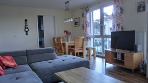 A living room with a grey sofa, a wooden coffee table, and a TV on a wooden stand. There is a dining area with a table and chairs, and a kitchen in the background. - 3-room apartment close to the beach for up to 6 people with elevator (Graal-Müritz)