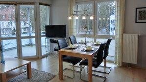 A dining area with a table set for four, a television on a stand, and large windows with curtains. - Strandnahe 2- Zi.-fewo für 3 Personen im 2. OG, mit Wlan (Graal-Müritz)