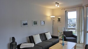 A modern living room with a dark grey sofa, a wooden coffee table, and a black armchair. There are three framed pictures on the wall, a tall floor lamp, and a window with curtains. - Strandnahe 2- Zi.-fewo für 3 Personen im 2. OG, mit Wlan (Graal-Müritz)