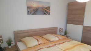 Een slaapkamer met een houten hoofdbord, een bed met een gebloemd plaid, een nachtkastje met een lamp, een potplant en een schilderij van een zonsondergang boven een strand.