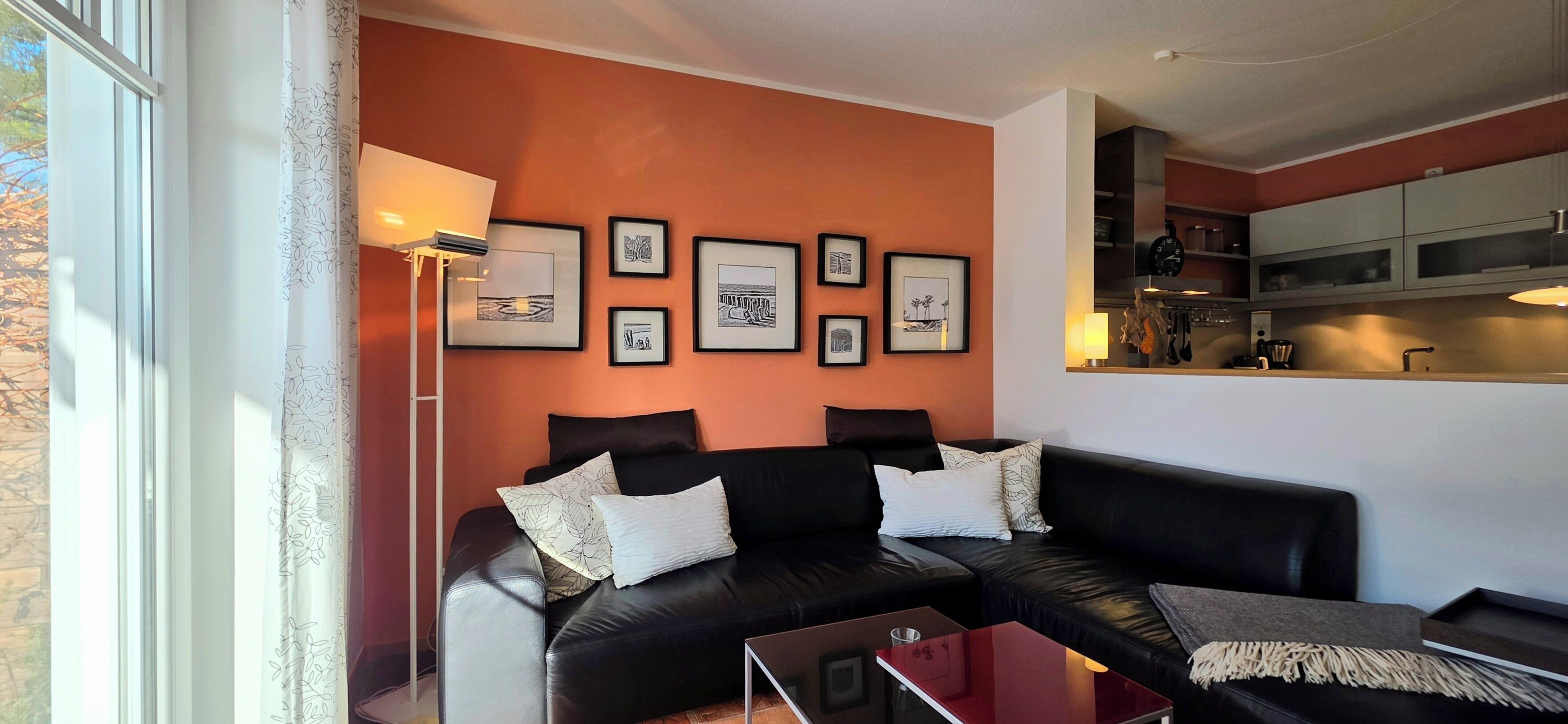 A living room with a black leather sofa, a glass coffee table, and a floor lamp. There are framed pictures on the wall and a kitchen area is visible in the background.