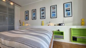 Une chambre moderne avec un grand lit, une porte-fenêtre coulissante et un mur décoré de tableaux encadrés et de petits vases jaunes.