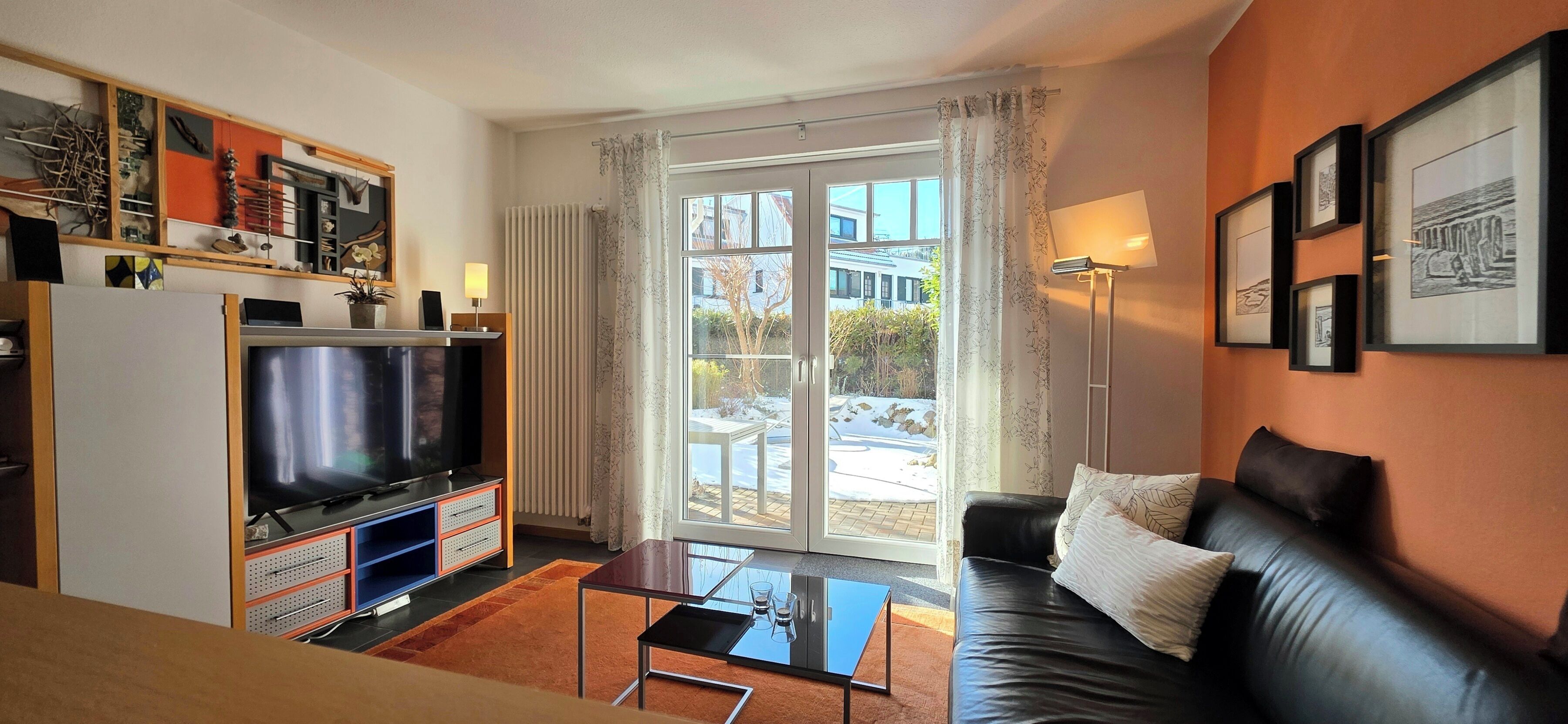 Ein Wohnzimmer mit Fernseher, Sofa, Couchtisch und Blick ins Freie durch ein großes Fenster.