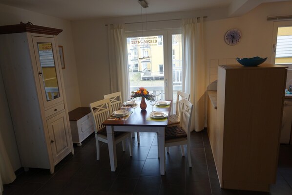 Een eetgedeelte met een tafel voor vier personen, witte stoelen, een vaas met bloemen en uitzicht op gebouwen door het raam.