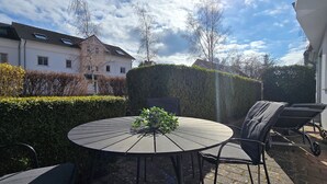 Une terrasse avec une table ronde et des chaises, un pot de fleurs et une haie.