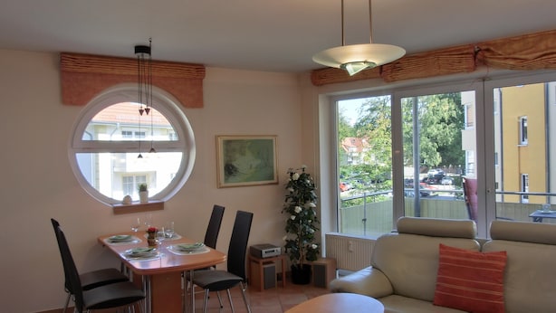 A dining area with a round table, chairs, and a window with a view of buildings and trees. - 3-zimmer-fewo im 1. OG mit Balkon mit 2 Schlafzimmern für 4 Personen, Wlan (Graal-Müritz)