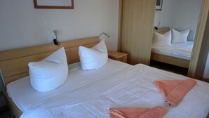 Una cama bien tendida con ropa de cama blanca y dos almohadas, un cabecero de madera, una mesita de noche con una lámpara y un espejo que refleja la habitación.