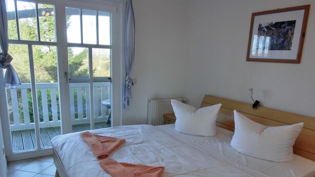 Habitación de hotel con cama, almohadas blancas, un cuadro enmarcado en la pared y vistas a un balcón con barandilla.