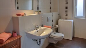 Un baño moderno con un lavabo blanco, espejo y retrete. Hay dos toallas rosas en la encimera y una pequeña planta en la estantería.