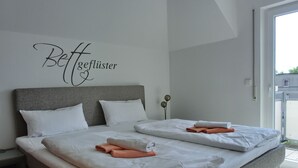 Una cama doble con sábanas blancas y toallas anaranjadas, un cabecero gris y un cartel en la pared que dice "Bettgeflüster".