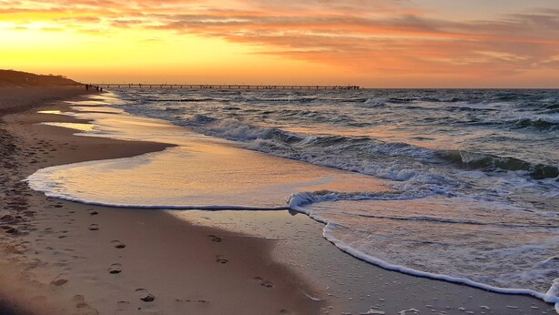 Ein Strand mit Wellen bei Sonnenuntergang, ein Pier in der Ferne und Fußabdrücke im Sand.