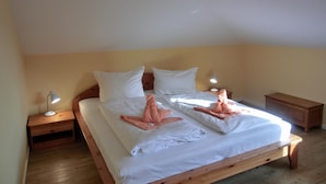 Una cama bien hecha con sábanas blancas y toallas rosas dispuestas sobre ella. Hay dos mesitas de noche con lámparas, y un mueble auxiliar de madera también con una lámpara. La habitación tiene suelo de madera y una pared amarilla lisa.