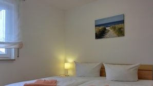 A double bed with white linens and two pink towels, a bedside lamp, and a painting of a beach scene on the wall.