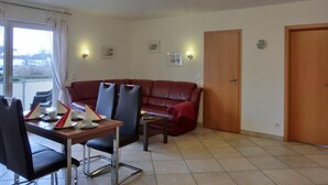 A living room with a red sofa, a wooden coffee table, and a dining area with chairs and a table set for two. There are wall-mounted lights and a door to the right.