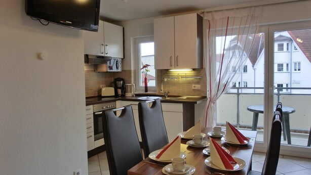 A modern kitchen with a flat-screen TV, a dining table set for four, and a balcony with a view of buildings.