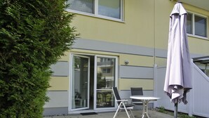 Een terras met een tafel en stoelen, een overdekte parasol en een glazen deur die naar de binnenruimte leidt.