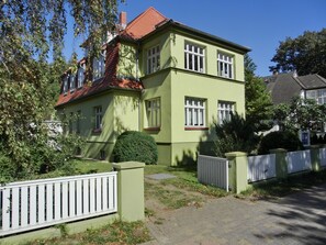 Une maison à deux étages, avec une façade verte, des encadrements de fenêtres blancs et un toit rouge.