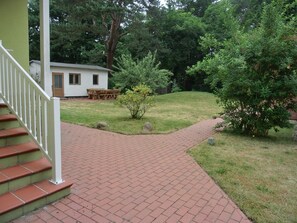 A brick pathway leading to a white building with a door and windows, surrounded by greenery and trees.