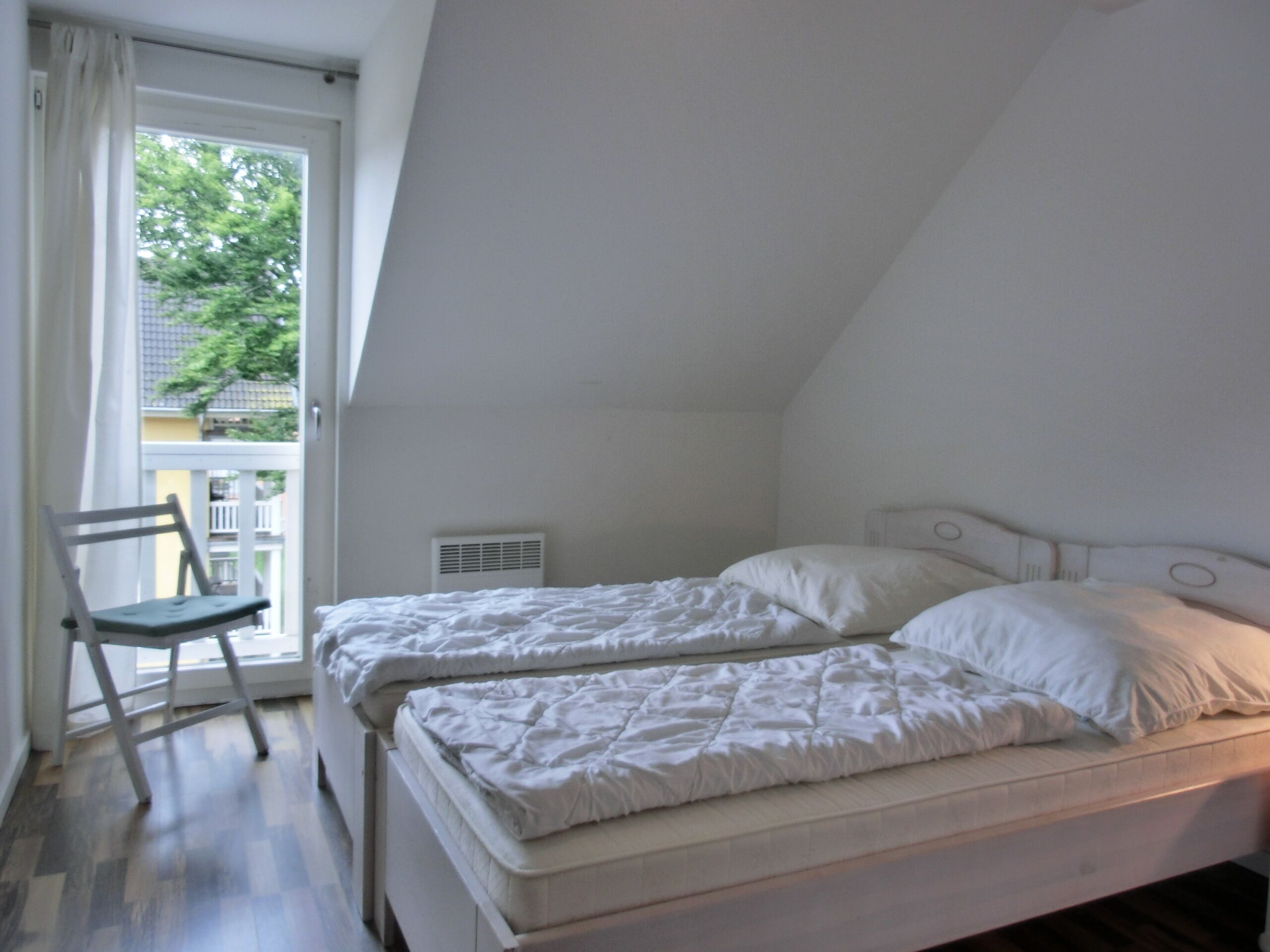 Un dormitorio con techo inclinado, una cama con ropa de cama blanca, una silla y una ventana con cortinas.