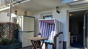 Un balcón con una mesa de madera y una silla a rayas, un cerramiento de celosía y una puerta que da a una habitación interior.