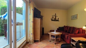 Ein gemütliches Wohnzimmer mit einem roten Sofa, einer Holzkommode, einem kleinen Tisch und einem schwarzen Ledersessel. Ein Fernseher ist an der Kommode befestigt, und rechts steht eine Lampe.