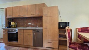 Eine kleine Küche mit Holzschränken, einem Geschirrspüler, einem Herd und einer Mikrowelle. Im angrenzenden Wohnbereich befindet sich ein kleiner Tisch mit einem Stuhl und eine Couch.