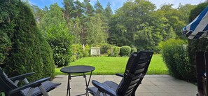 Une terrasse avec deux chaises et une petite table ronde, entourée d’une végétation luxuriante et d’une pelouse bien entretenue.