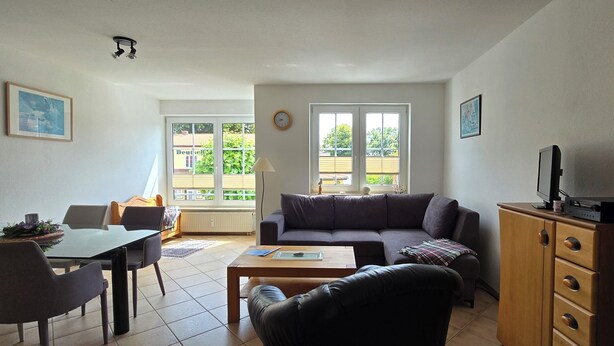 A living room with a sofa, coffee table, and dining area with chairs. There are windows, a clock, and a TV on a cabinet.