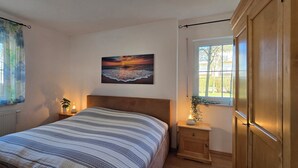 Ein Schlafzimmer mit einem Bett, einem Nachttisch mit Lampe, einem Fenster mit Vorhängen und einem Sonnenuntergangsbild.
