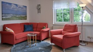 A living room with a red sofa, a glass coffee table, and a red armchair. There is a painting on the wall, a window with a view of greenery, and a lamp. - 3-zi.-fewo mit 2 Schlafzimmern + Kostenfreien W-lan-zugang (Graal-Müritz)