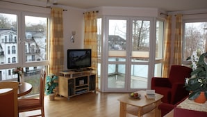 A living room with a television, a dining table, and chairs. There is a large window with curtains, and a plant is visible. - Strandnahe 3-zimmer-fewo im 3.og für 4 Pers. mit Wlan (Graal-Müritz)