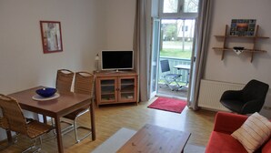 A living room with a wooden dining table, wicker chairs, a red sofa, a TV on a wooden cabinet, and a view of the outdoors through a glass door. - 2-zi-fewo für 4 Pers. mit Wlan, Haustiere Erlaubt (Graal-Müritz)