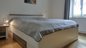 A bedroom with a bed, a nightstand, a lamp, a window with curtains, and a painting on the wall. - 2-zi-fewo für 4 Pers. mit Wlan, Haustiere Erlaubt (Graal-Müritz)