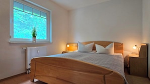 A bedroom with a wooden bed, two bedside lamps, a window with a view of greenery, and a radiator. - 2-zimmer-fewo für 3 Pers. Inkl. Strandkorb (Graal-Müritz)