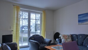 Een zithoek met een bank, fauteuil en een klein tafeltje. Er is een raam met gordijnen, een radiator en een schilderij aan de muur.
