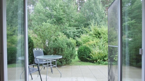 Porta de vidro que dá para um alpendre com mesa e cadeiras, rodeado por muita vegetação.