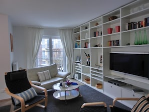 Uma sala de estar moderna com sofá, poltronas, mesa de centro e estante repleta de livros e objetos decorativos.