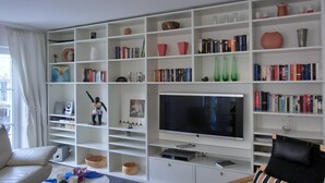 Una sala de estar con un televisor de pantalla plana grande, estanterías blancas con libros y objetos decorativos, un sofá y una mesa de centro.